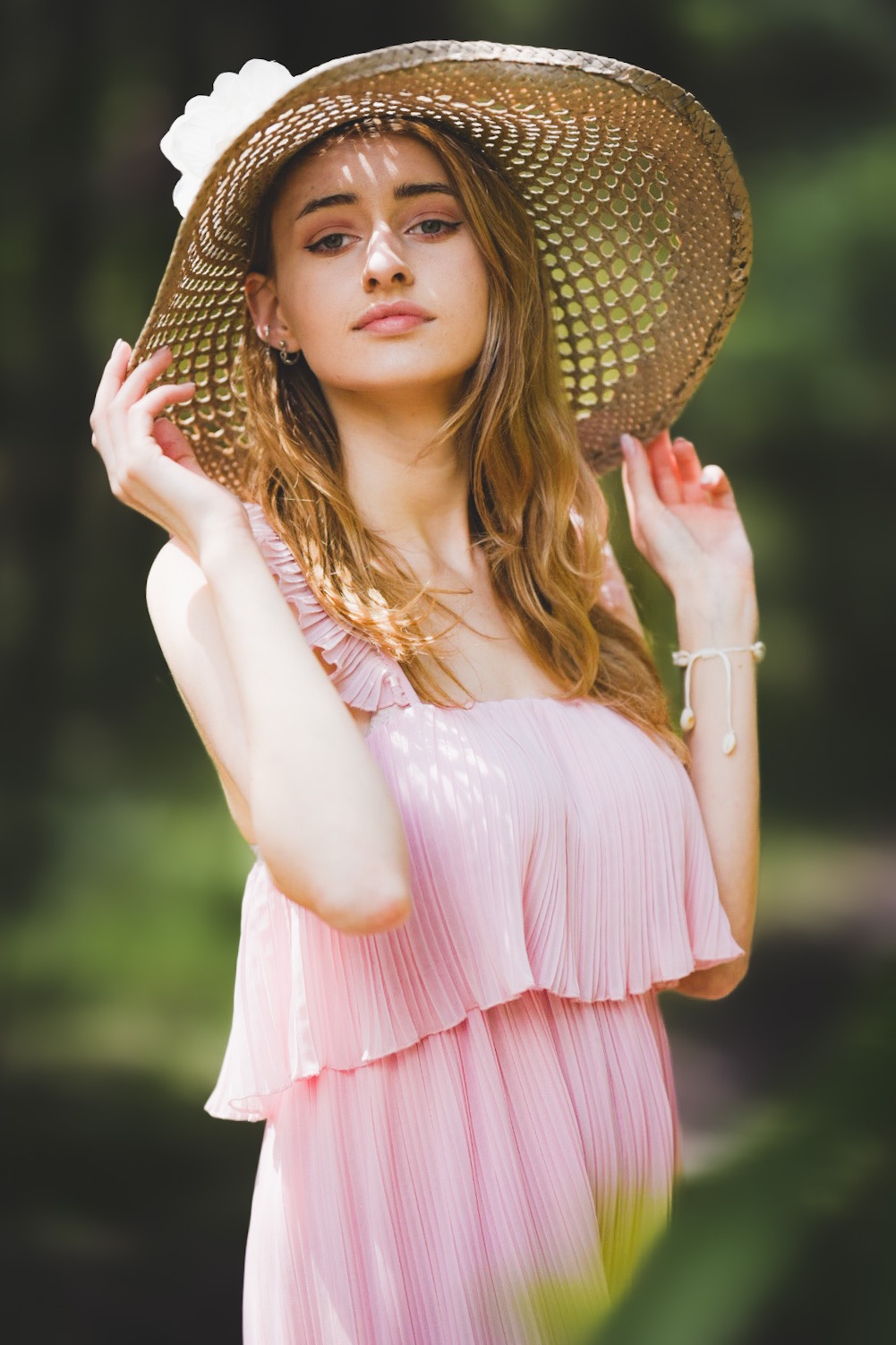 Portret kobiety w różowej sukience i słomkowym kapeluszu na tle zieleni, z delikatnym uśmiechem i naturalnym makijażem.