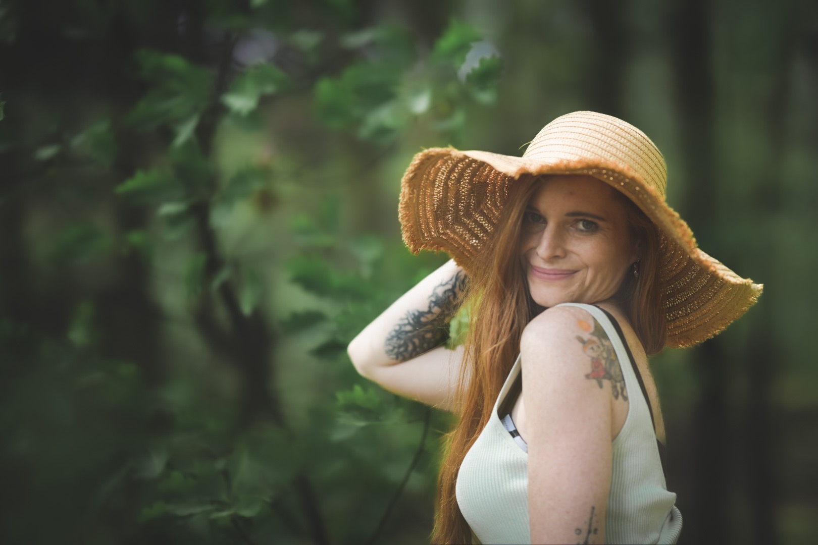 Portret kobiety w słomkowym kapeluszu z szerokim rondem, z tatuażami na ramionach, na tle zielonych liści