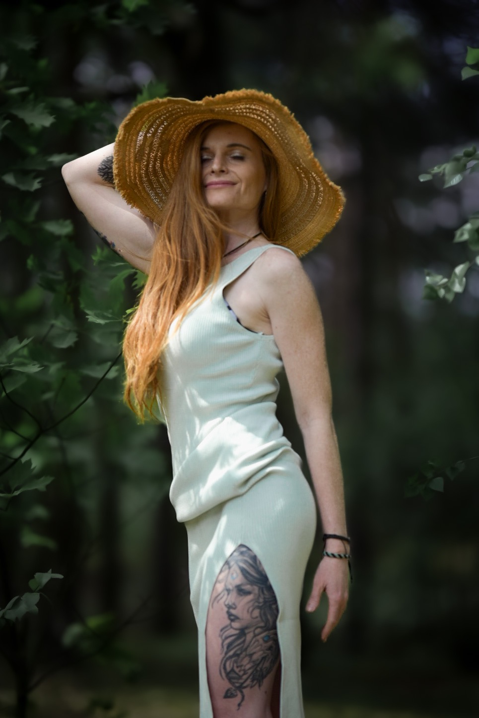 Kobieta z rudymi włosami w słomkowym kapeluszu i jasnej sukience z rozcięciem, eksponującym tatuaż na udzie, pozuje na tle zieleni.