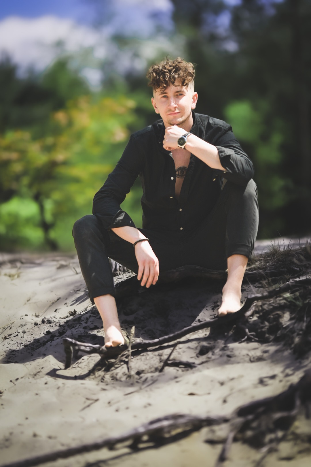 Młody mężczyzna z tatuażem na piersi, ubrany na czarno, siedzi boso na piaszczystym wzgórzu z korzeniami drzew w tle.