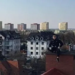 Kominiarz w pełnym stroju siedzi na kominie na tle miejskiego krajobrazu z blokami mieszkalnymi w oddali.