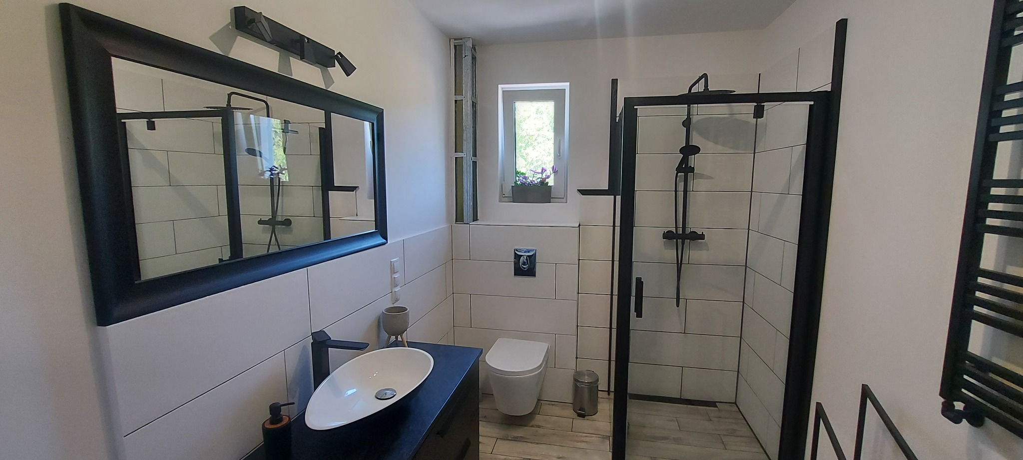 Nowoczesna łazienka z białą armaturą, czarną kabiną prysznicową typu walk-in i owalną umywalką nablatową, oświetlona światłem dziennym z okna z kwiatami.