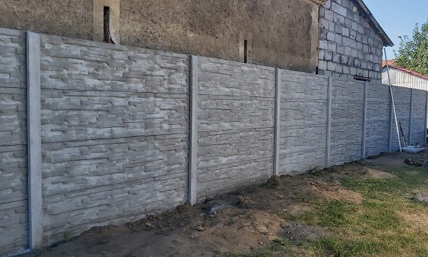 Szary, betonowy płot panelowy o fakturze kamienia, oddzielający posesję od sąsiada, widoczny fragment budynku w tle.