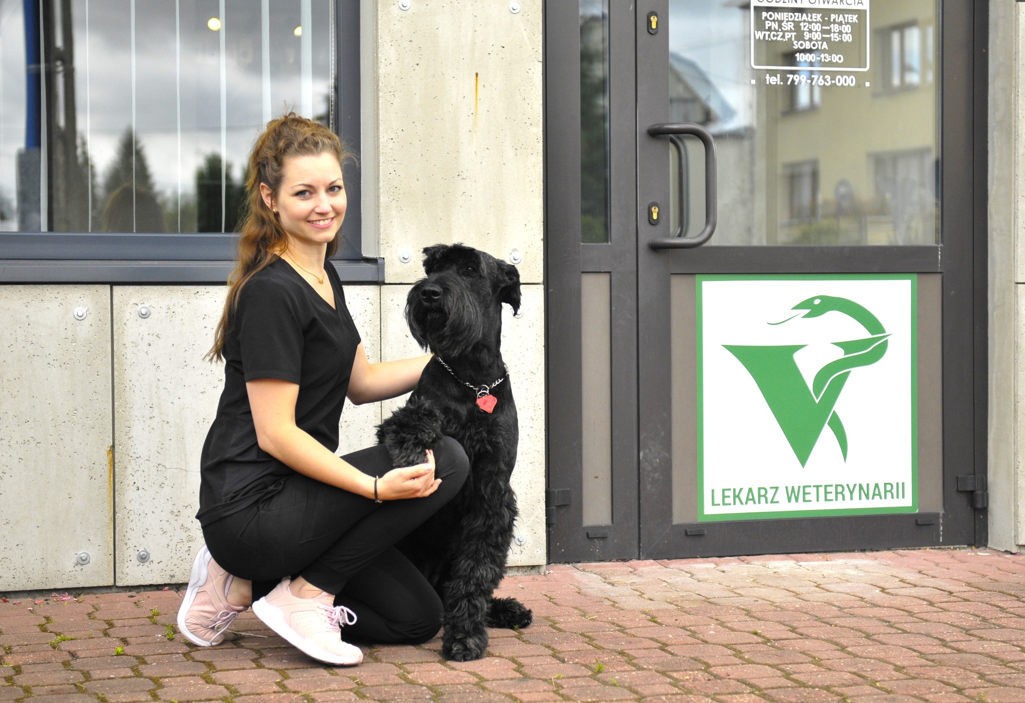 Kobieta w czarnym stroju kuca obok czarnego sznaucera olbrzyma przed wejściem do gabinetu, na drzwiach logo z napisem: Lekarz Weterynarii.