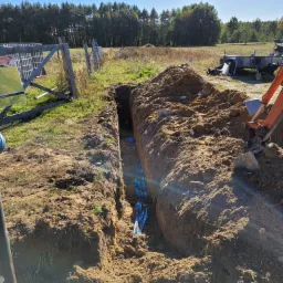 Wąski wykop w ziemi z ułożonymi niebieskimi rurami, widoczny fragment koparki i przyczepy w tle, słoneczny dzień.