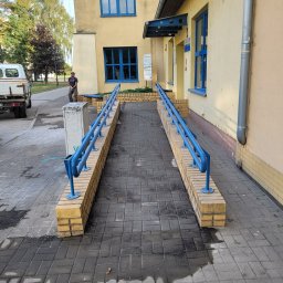 Usługi budowlano sprzętowe Mariusz Rosiński - Rampa dla niepełnosprawnych z niebieskimi poręczami prowadząca do budynku o żółtej elewacji i niebieskich oknach. Widoczny fragment chodnika z kostki brukowej.