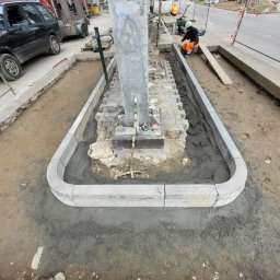 Usługi budowlano sprzętowe Mariusz Rosiński - Montaż obrzeży betonowych wokół słupa oświetleniowego, widoczny pracownik w stroju roboczym i narzędzia. Etap prac ziemnych i betonowych na zewnątrz.