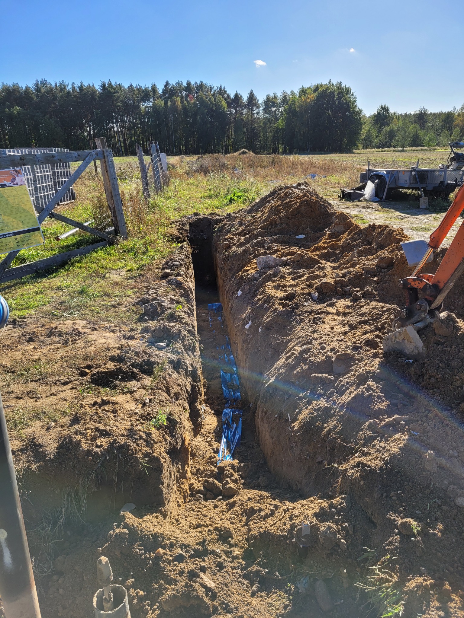 Wąski wykop w ziemi z ułożonymi niebieskimi rurami, widoczny fragment koparki i przyczepy w tle, słoneczny dzień.