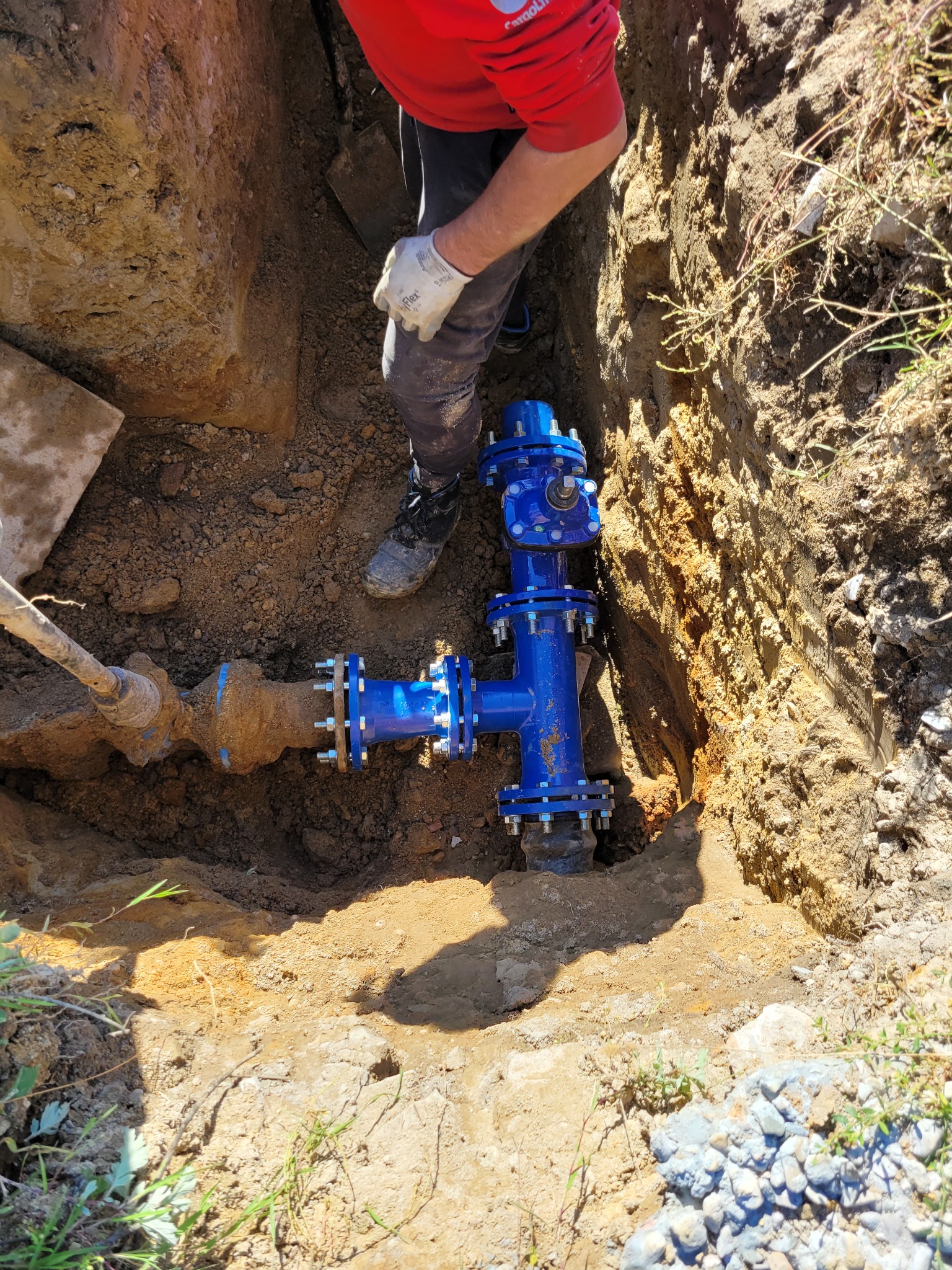 Niebieska armatura wodociągowa z kołnierzami w wykopie ziemnym, widoczny fragment starej, zardzewiałej rury, pracownik w czerwonej koszulce i białych rękawicach nad wykopem.