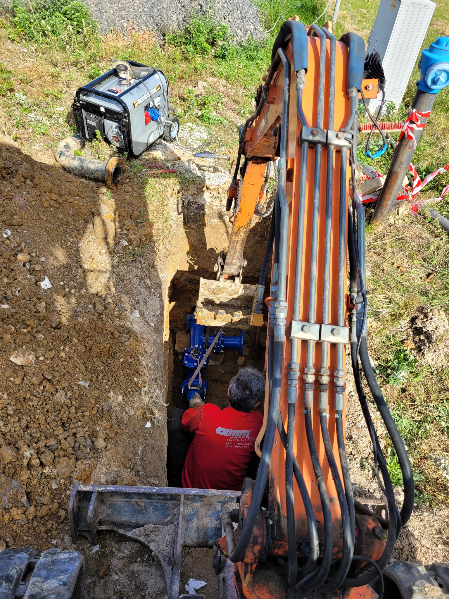 Pracownik w czerwonej koszulce w głębokim wykopie instaluje niebieskie rury przyłącza wodno-kanalizacyjnego, widoczny pomarańczowy wysięgnik koparki i agregat prądotwórczy na zewnątrz wykopu.