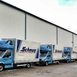 Flota błękitnych samochodów dostawczych z naczepami, oznaczonych logo firmy transportowej, zaparkowana w rzędzie przed magazynem z widoczną wieżą telekomunikacyjną w tle.