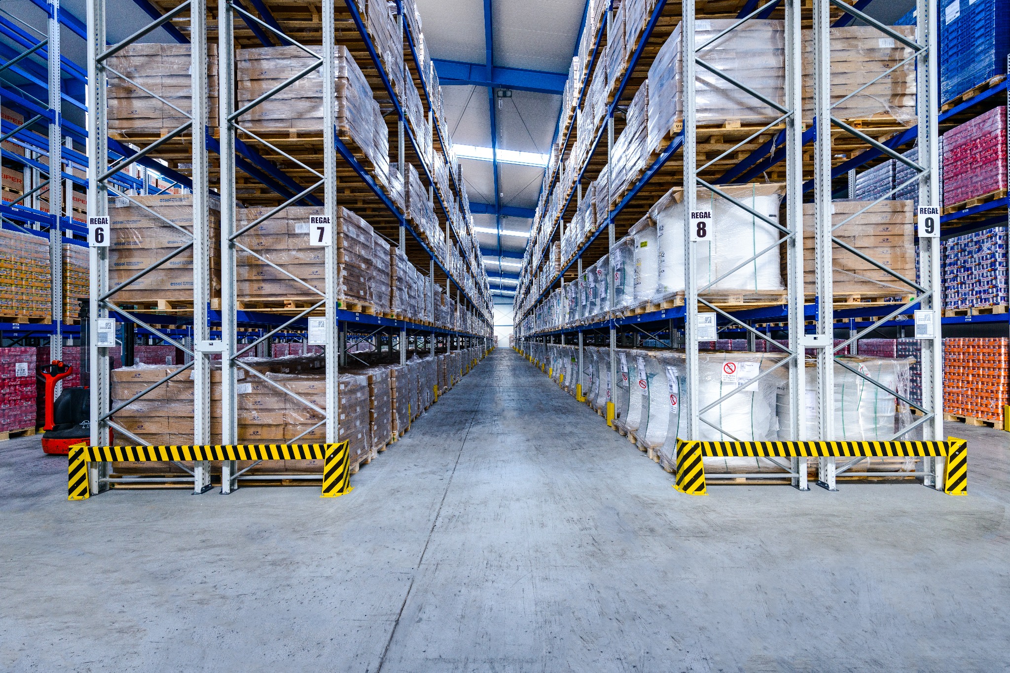 Pełny magazyn z wysokimi regałami wypełnionymi paletami zafoliowanych towarów, widoczne numery regałów i żółto-czarne zabezpieczenia na podłodze.