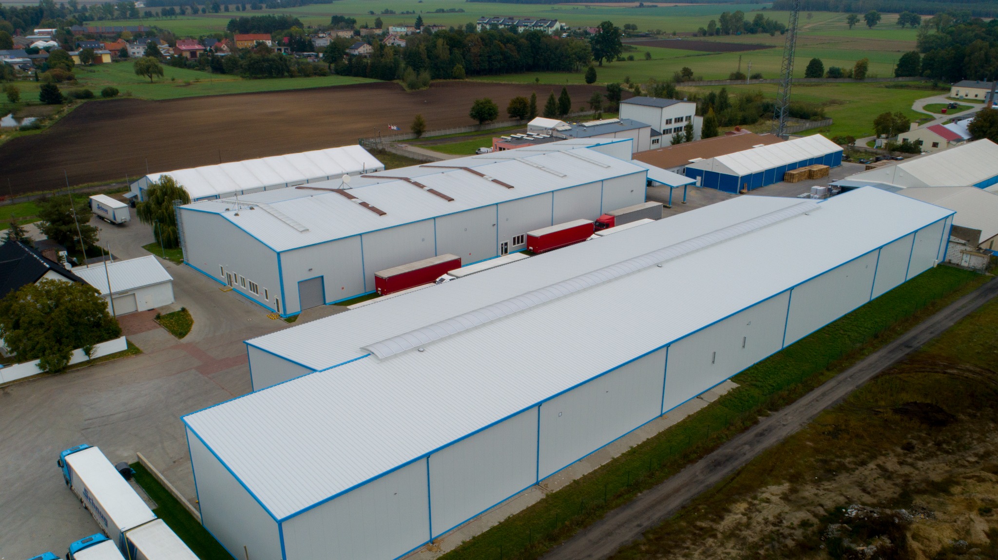 Widok z lotu ptaka na kompleks magazynowy z białymi budynkami i niebieskimi akcentami, z czerwonymi naczepami ciężarówek zaparkowanymi w pobliżu.
