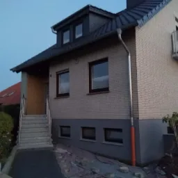 Dwukondygnacyjny dom z cegły klinkierowej z szarym dachem, widoczna ocieplona i pomalowana podmurówka oraz przygotowane podłoże pod kostkę brukową.