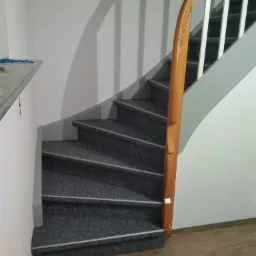 Wewnętrzne schody z szarym dywanem na stopniach, drewniana poręcz po prawej stronie i białe ściany, widok z dołu na pierwszy bieg schodów.