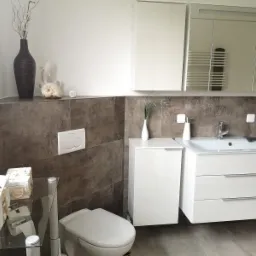 Nowoczesna łazienka z brązowymi płytkami imitującymi kamień, białą armaturą, wiszącą szafką z umywalką i lustrzaną szafką nad nią, dekoracje w postaci wazonu z suchymi gałązkami i muszli.