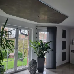 Nowoczesne wnętrze z dekoracyjnym sufitem w odcieniach szarości, oświetlonym od spodu, duże okna z widokiem na ogród, rośliny doniczkowe i błyszcząca podłoga z płytek.