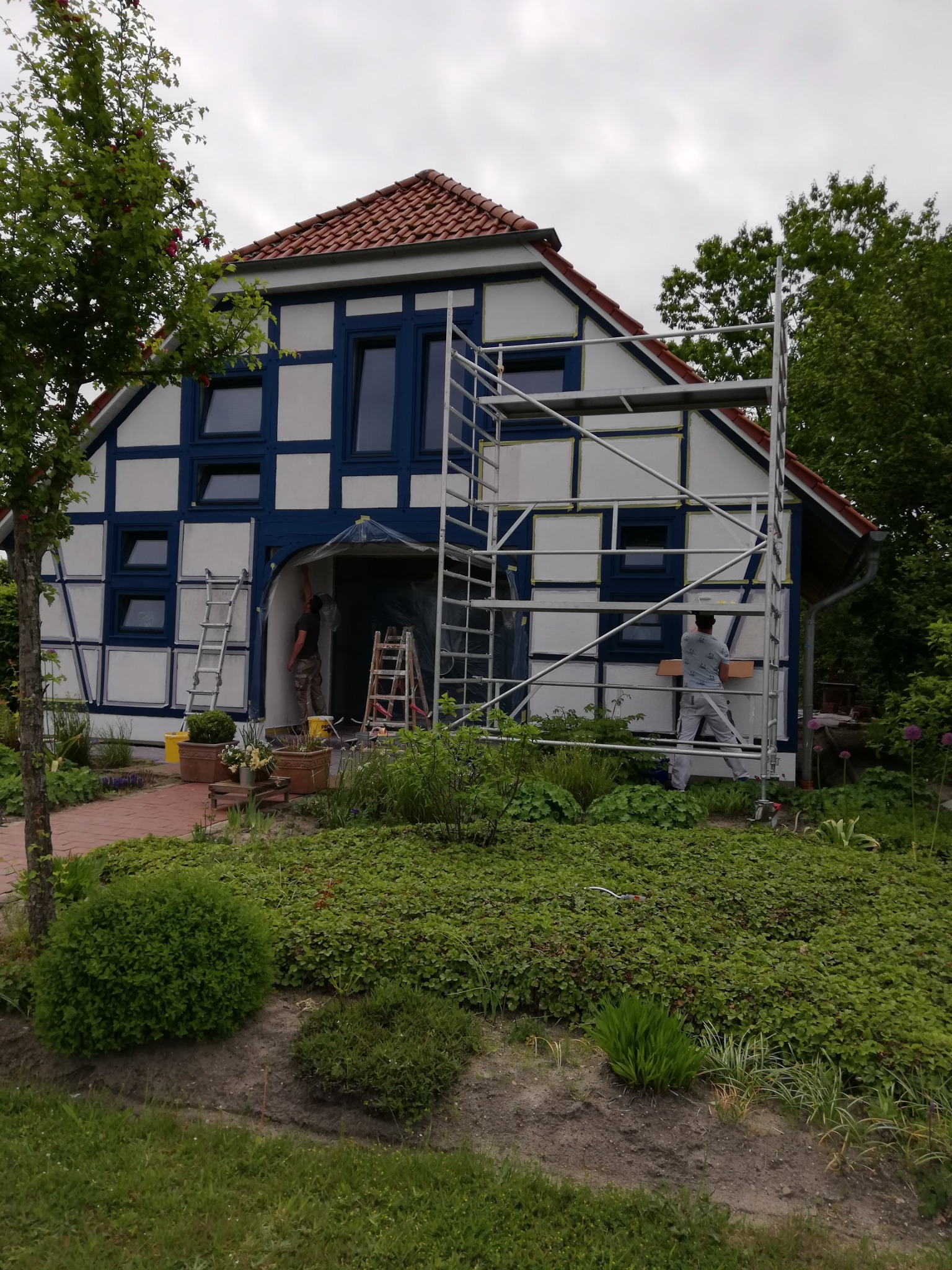 Dom szachulcowy w trakcie renowacji elewacji, widoczne rusztowania, zabezpieczone okna taśmą malarską, pracownicy w trakcie pracy, ogród z bujną roślinnością.