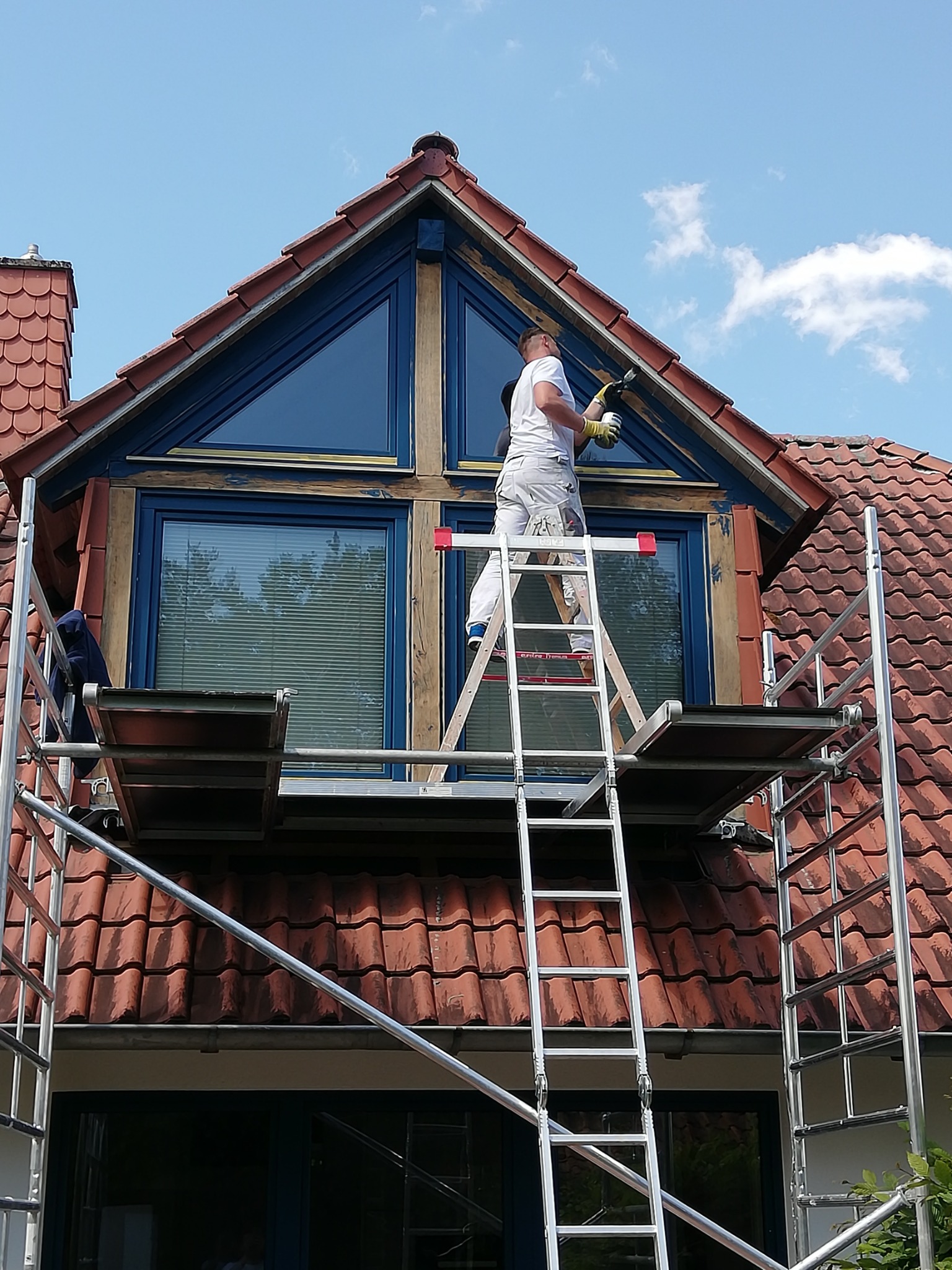 Pracownik na rusztowaniu maluje ramę okienną na poddaszu domu z czerwoną dachówką, widoczne niedomalowane fragmenty i pędzel w dłoni.