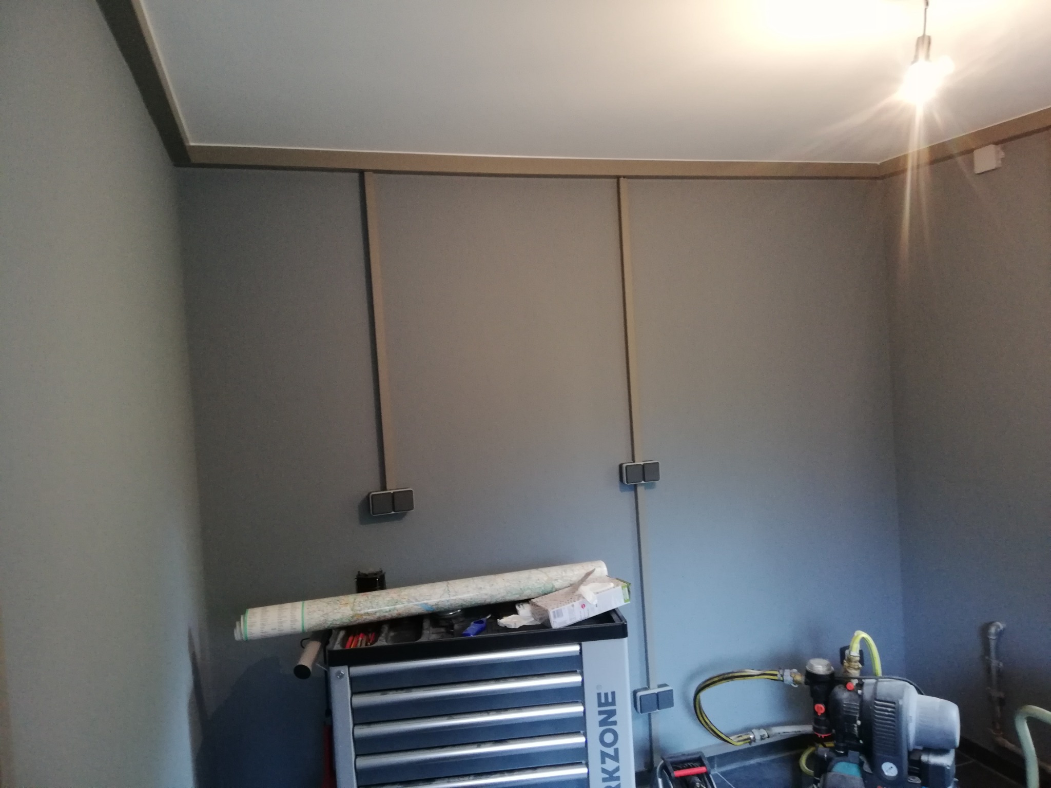 Szara ściana z widocznymi przewodami elektrycznymi w listwach, narzędzia i maszyna na podłodze, sufit wykończony listwami dekoracyjnymi.