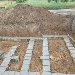 Wylane fundamenty z szalunkiem drewnianym w wykopie, widoczna sterta ziemi i krajobraz w tle.