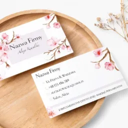 Dwie wizytówki z motywem kwitnącej wiśni, leżące na drewnianej tacy obok gałązki suszonych kwiatów. Wizytówki prezentują dane firmy i osoby, z adresem w Warszawie.