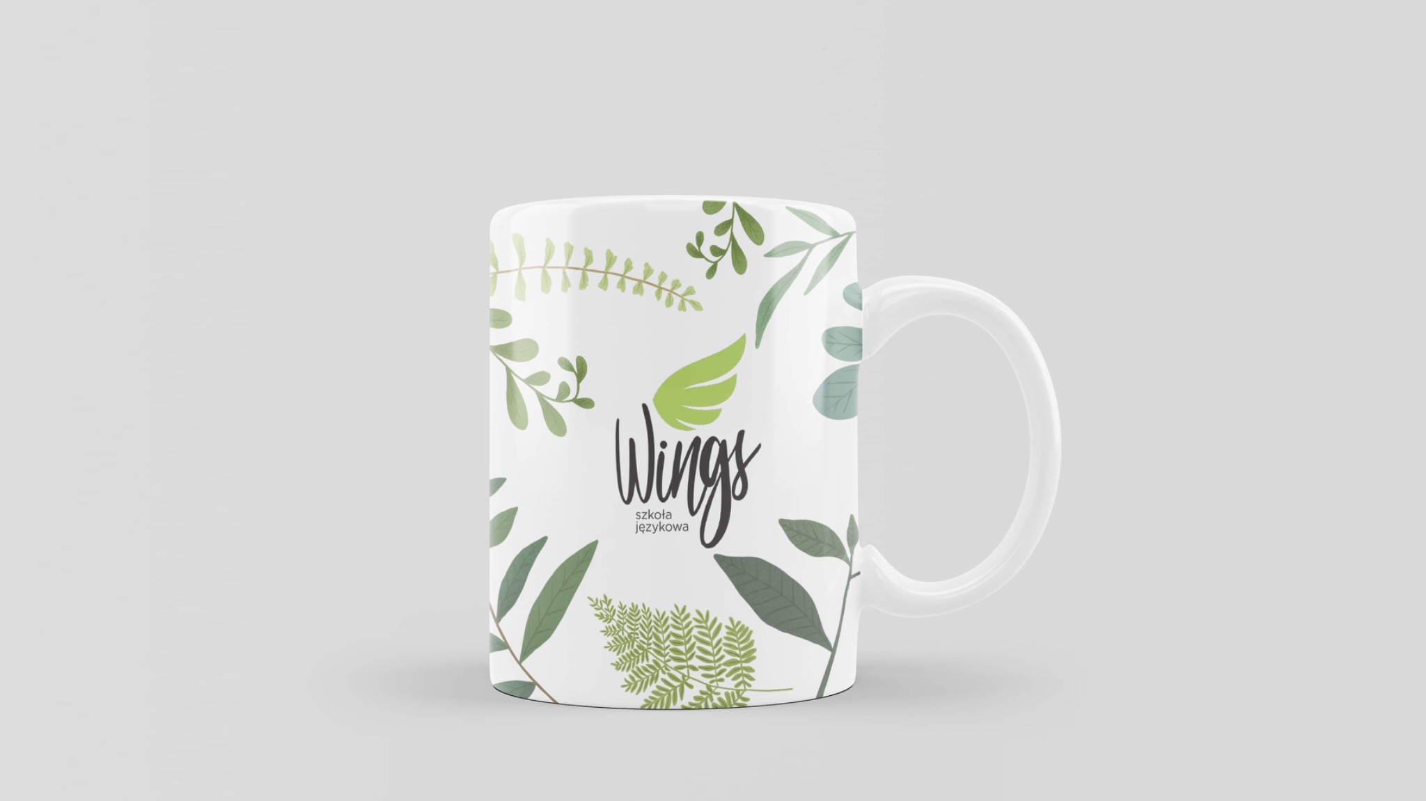 Biały kubek z motywem roślinnym i logo szkoły językowej Wings na jasnoszarym tle.