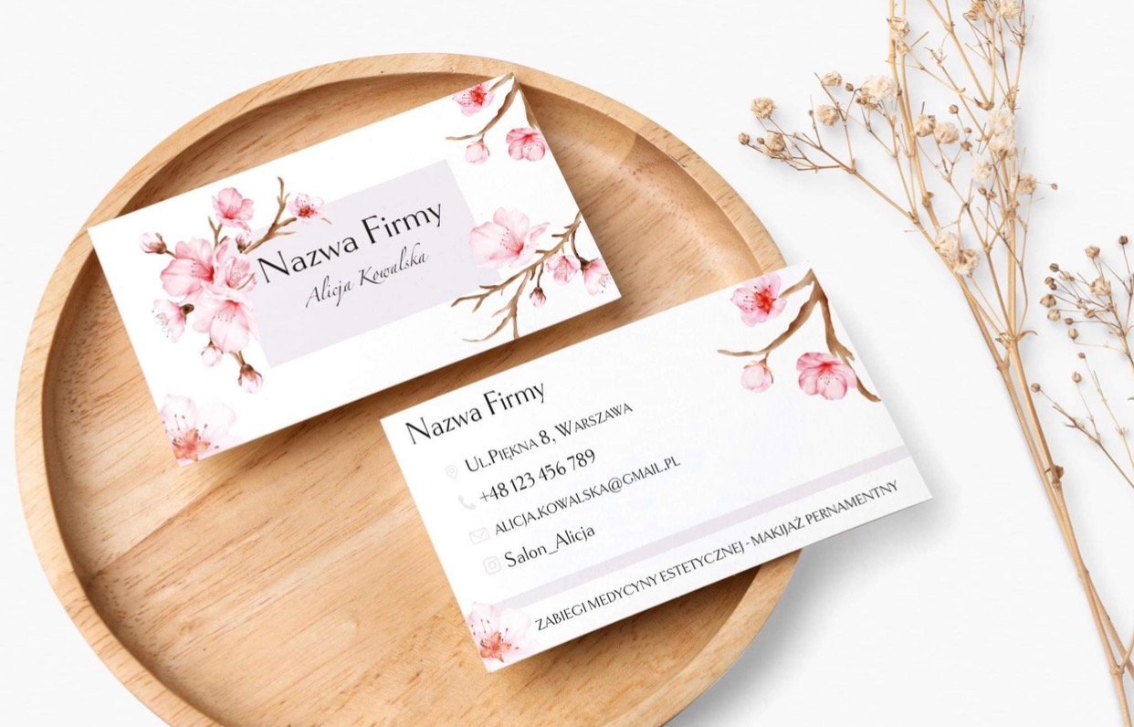 Dwie wizytówki z motywem kwitnącej wiśni, leżące na drewnianej tacy obok gałązki suszonych kwiatów. Wizytówki prezentują dane firmy i osoby, z adresem w Warszawie.