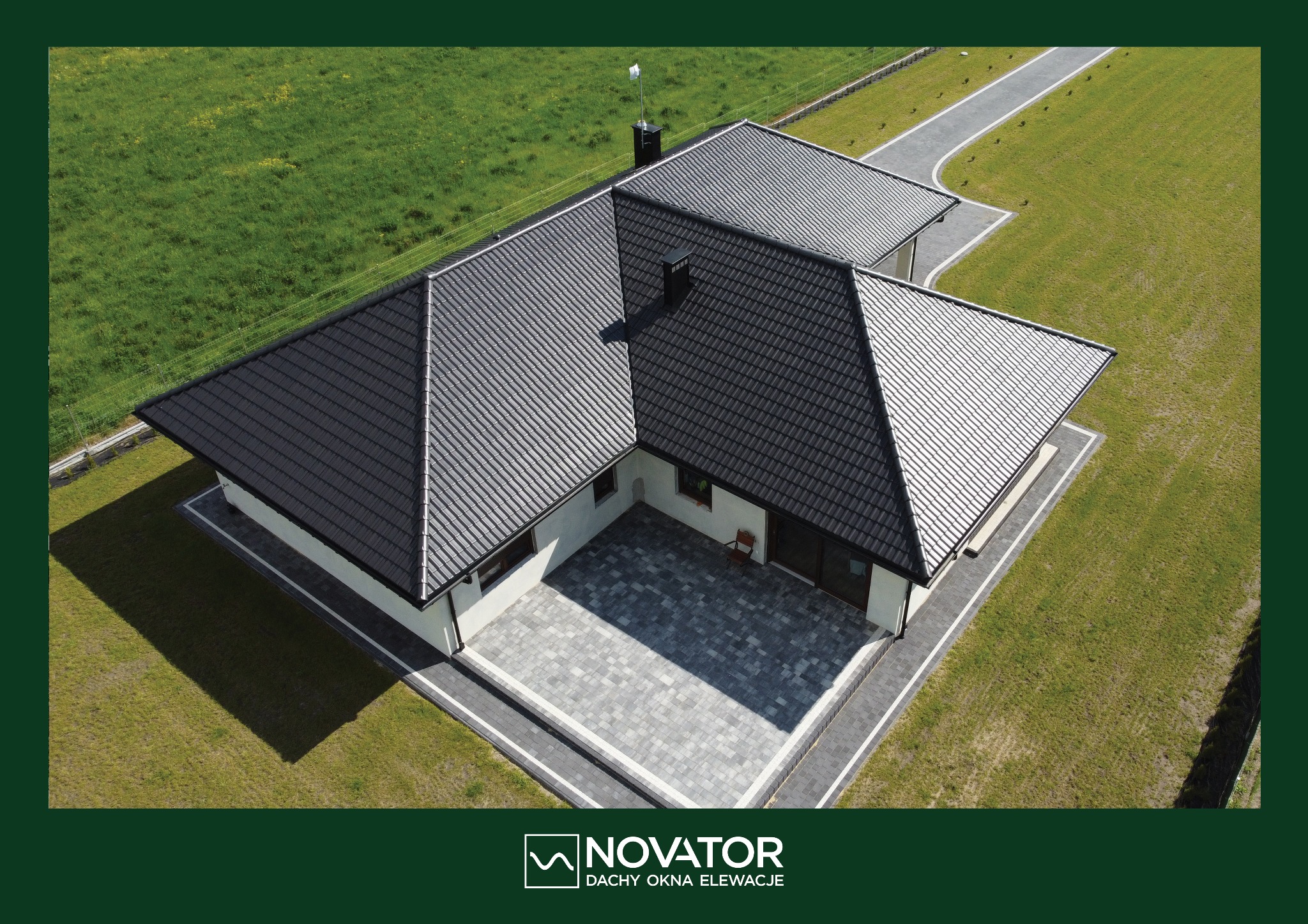 Nowoczesny dom jednorodzinny z ciemnym dachem widziany z lotu ptaka, otoczony zielonym trawnikiem i szarym brukowanym patio.