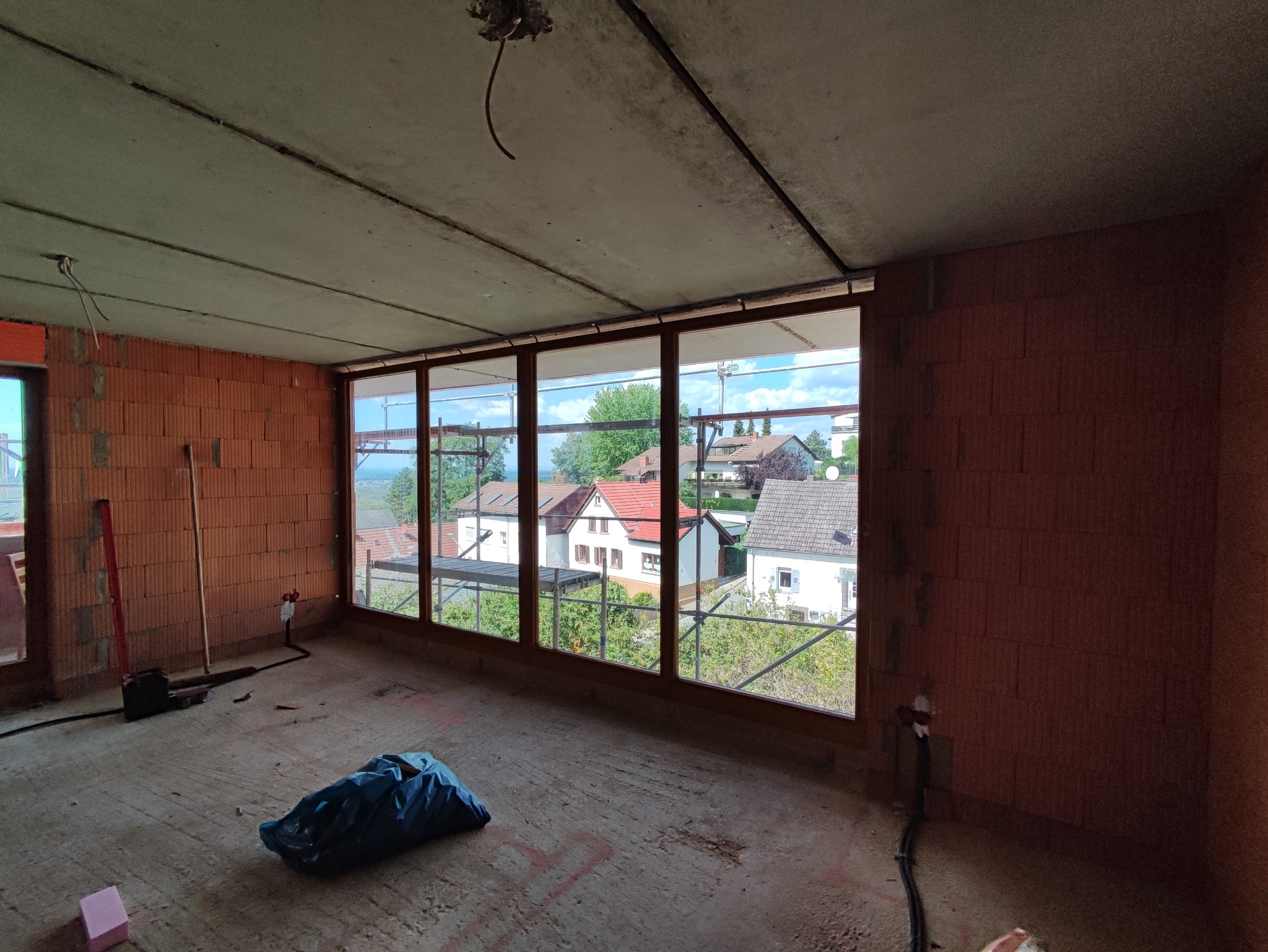 Wnętrze surowego budynku z widokiem na domy przez duże okno, widoczne ceglane ściany, betonowy sufit, narzędzia i worek na podłodze.