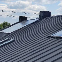 Nowy, grafitowy dach z blachy trapezowej z zamontowanymi oknami dachowymi i świetlikami, widok z dołu na tle błękitnego nieba z chmurami.