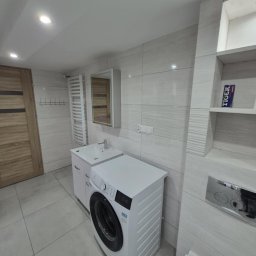 DAMSEB - Jasna łazienka po remoncie z pralką, umywalką z szafką, grzejnikiem drabinkowym i płytkami imitującymi drewno na ścianach. Widoczna toaleta i drewniane drzwi.