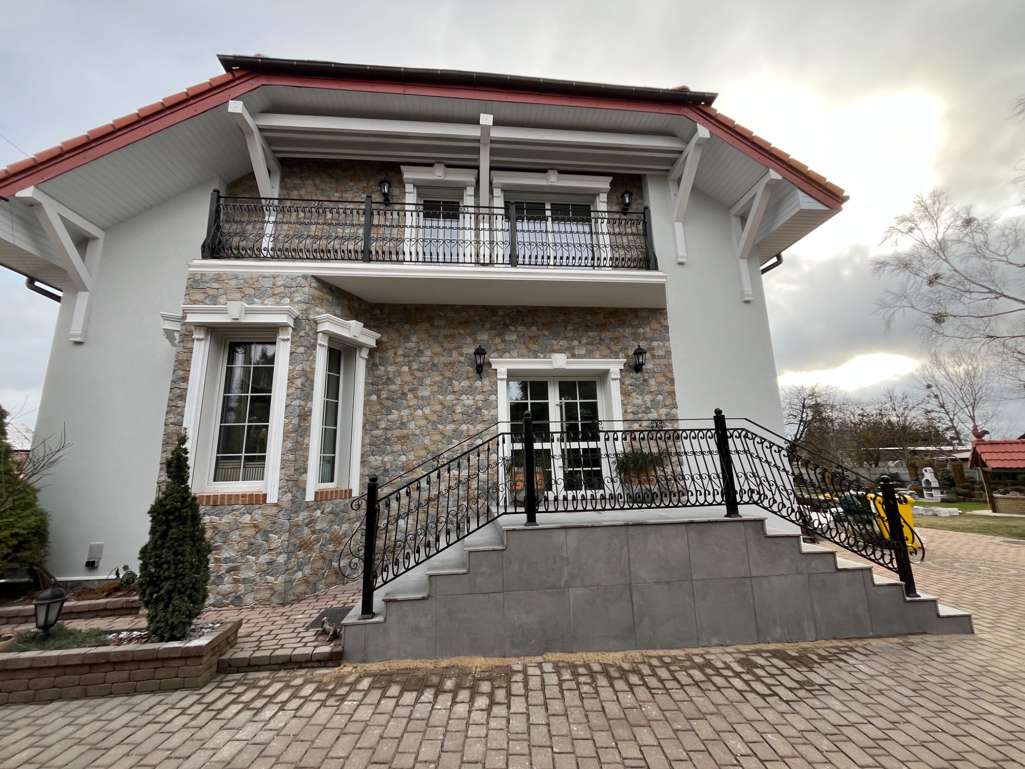 Dwupiętrowy dom z kamienną elewacją, balkonem z kutą balustradą i schodami wejściowymi. Widoczne detale wykończeniowe okien i dachu. Brukowany podjazd.
