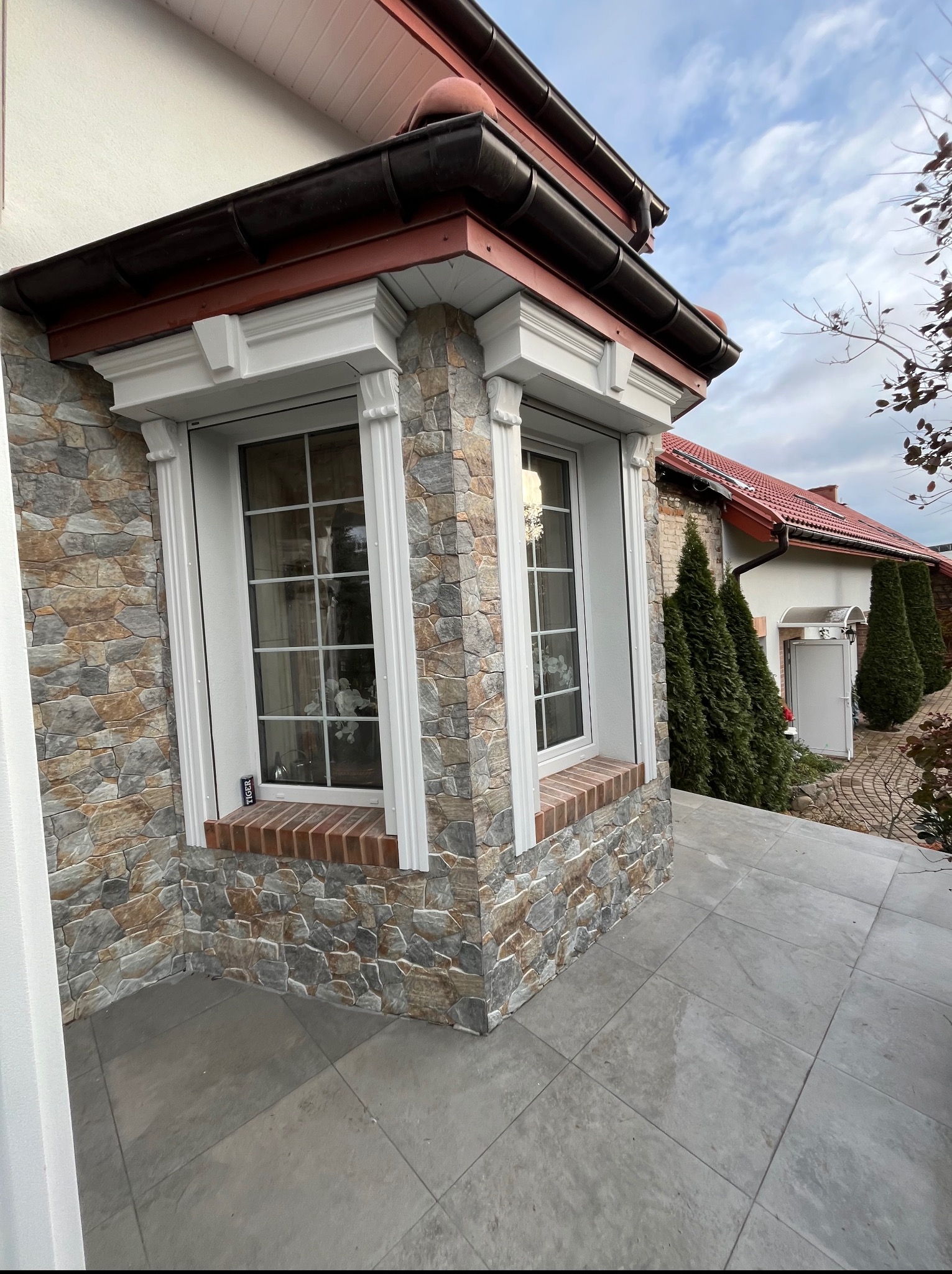 Eleganckie okno z białymi ramami i dekoracyjnymi kolumnami, osadzone w kamiennej elewacji. Widoczny fragment tarasu z szarych płyt i fragment budynku w tle.