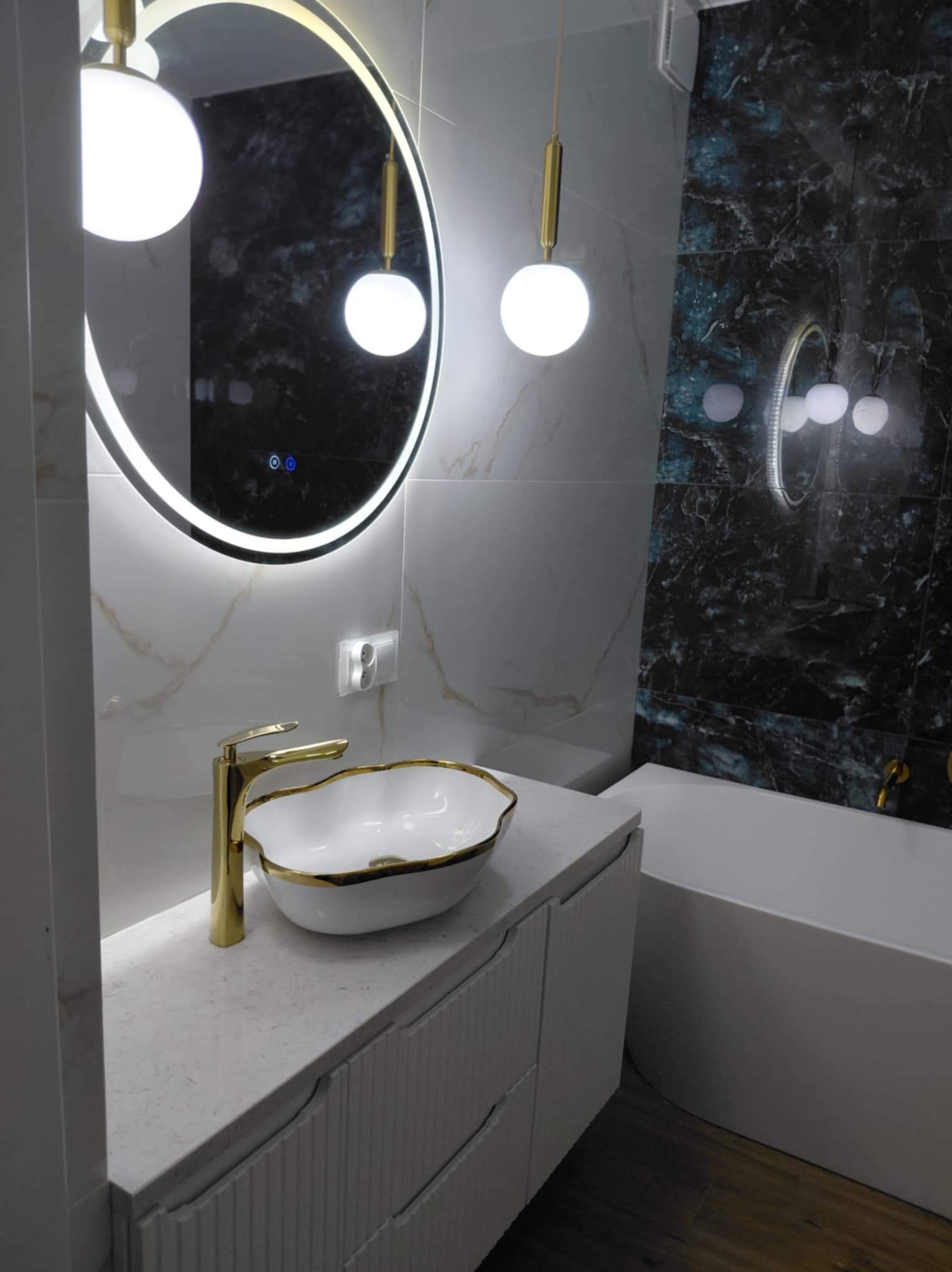 Elegancka łazienka z okrągłym lustrem LED, złotą armaturą i umywalką nablatową. Marmurowe płytki w odcieniach bieli i czerni. Nowoczesne oświetlenie.