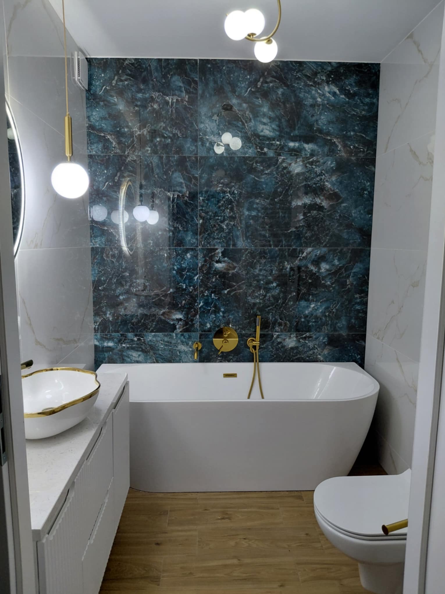 Nowoczesna łazienka z wanną, umywalką na blacie i toaletą. Ciemne płytki imitujące marmur na ścianie, złote detale armatury i oświetlenia, drewnopodobna podłoga.