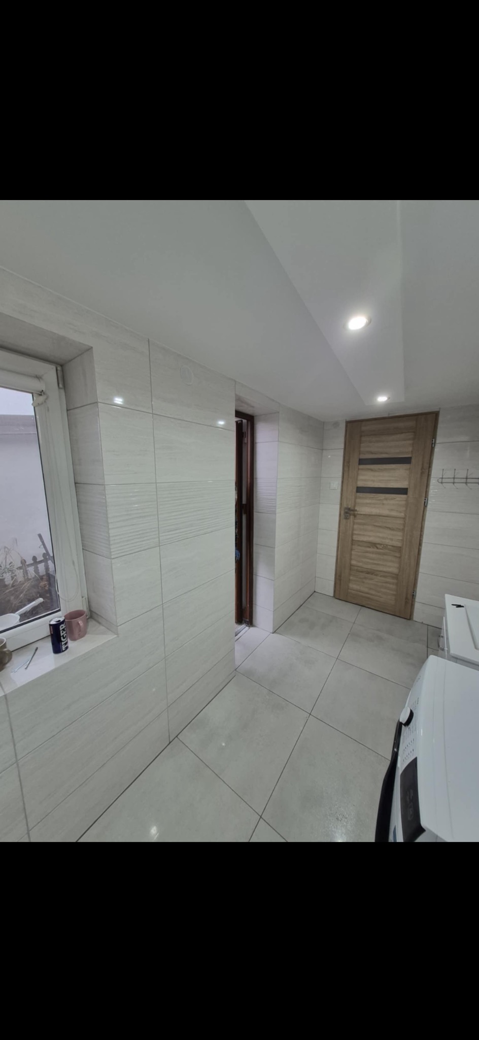 Jasne płytki ścienne i podłogowe w korytarzu z oknem, drewnianymi drzwiami i widoczną pralką. Minimalistyczny design, świeżo po remoncie.