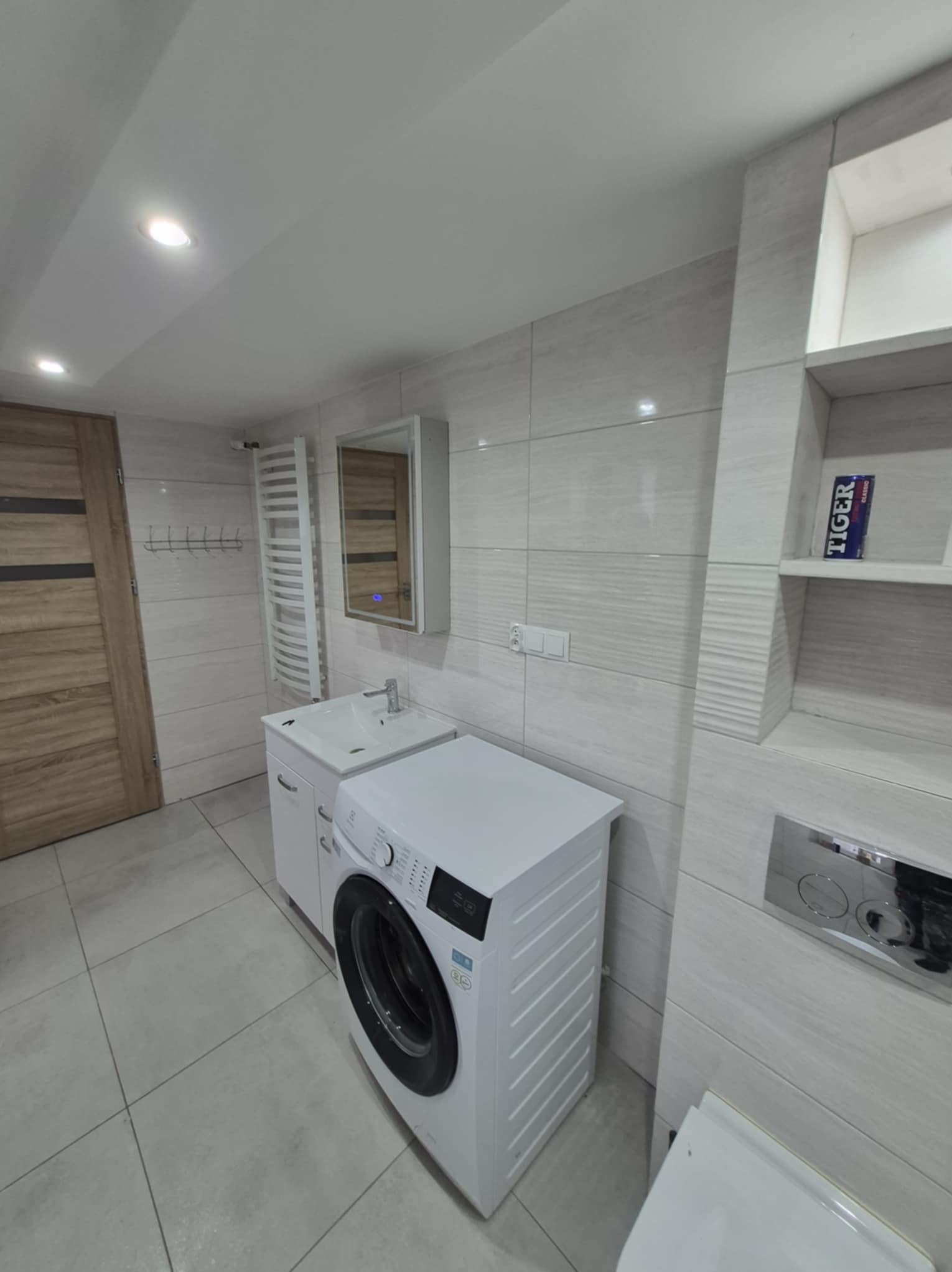 Jasna łazienka po remoncie z pralką, umywalką z szafką, grzejnikiem drabinkowym i płytkami imitującymi drewno na ścianach. Widoczna toaleta i drewniane drzwi.