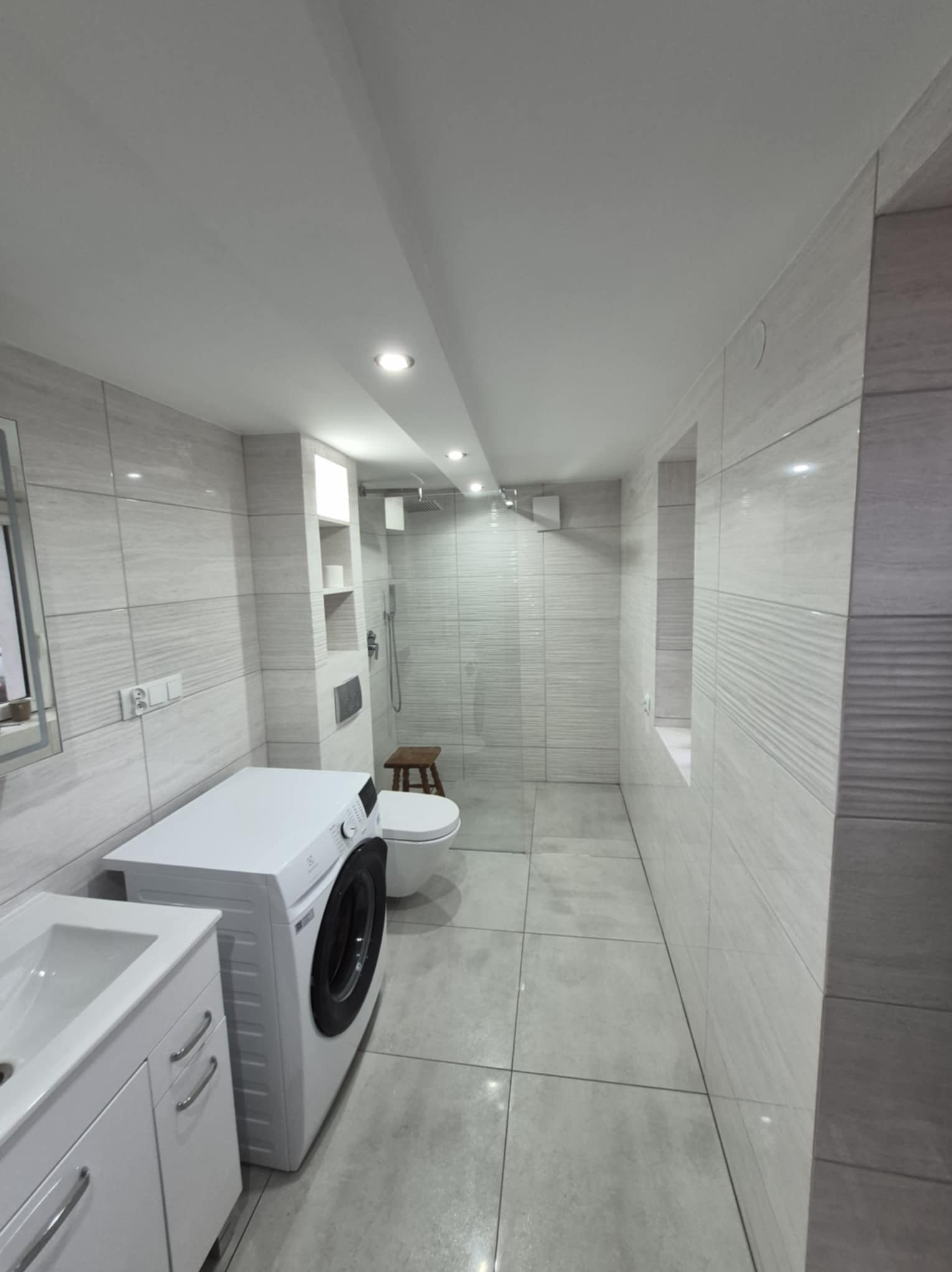 Jasna łazienka po remoncie z pralką, umywalką z szafką, toaletą i kabiną prysznicową. Ściany i podłoga wyłożone płytkami w odcieniach szarości. Minimalistyczny styl.