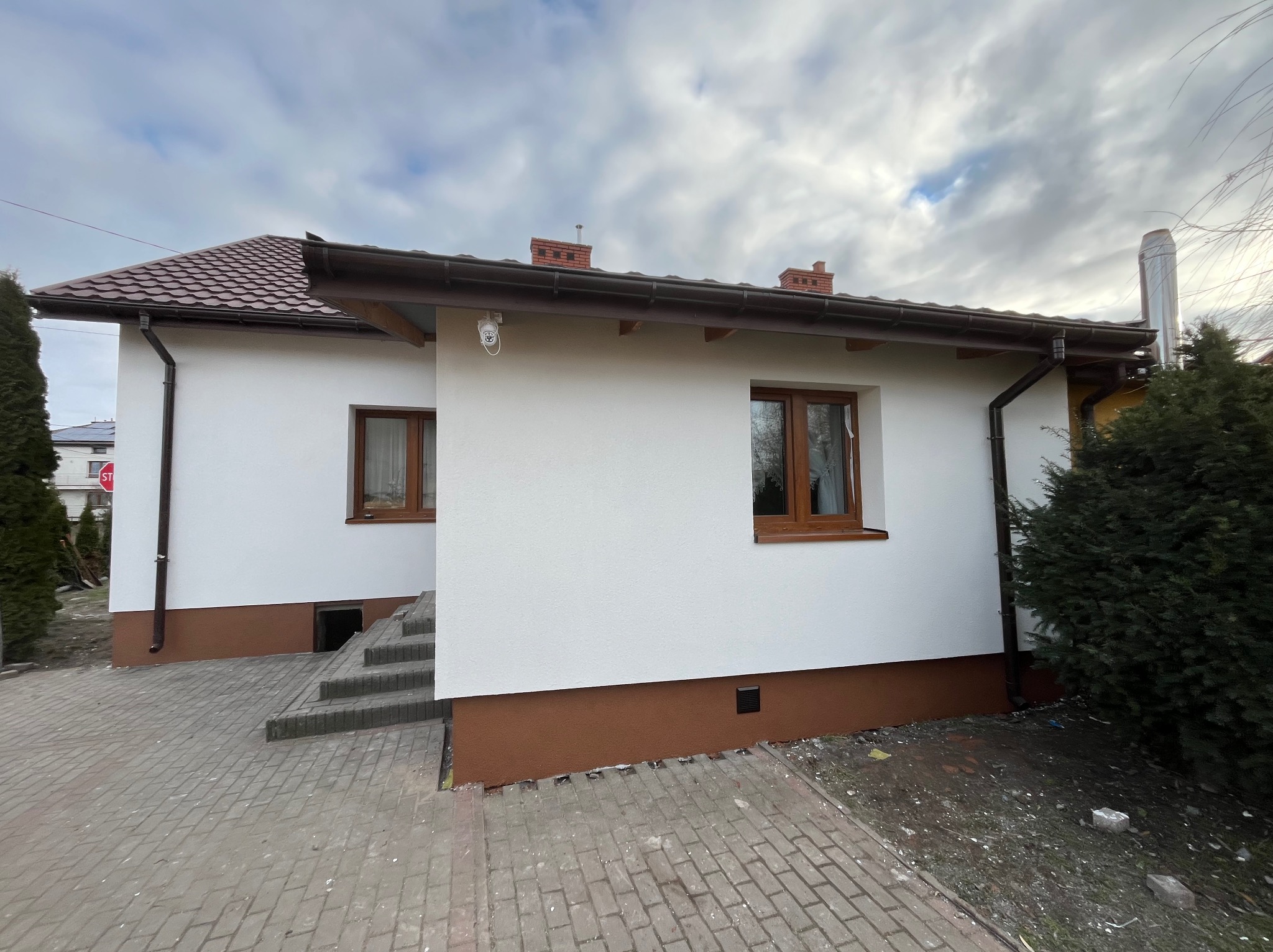 Biały dom z brązowymi oknami i dachem, świeżo po remoncie elewacji. Widoczny fragment chodnika z kostki brukowej i fragment ogrodu. Ujęcie z dołu, pochmurne niebo.