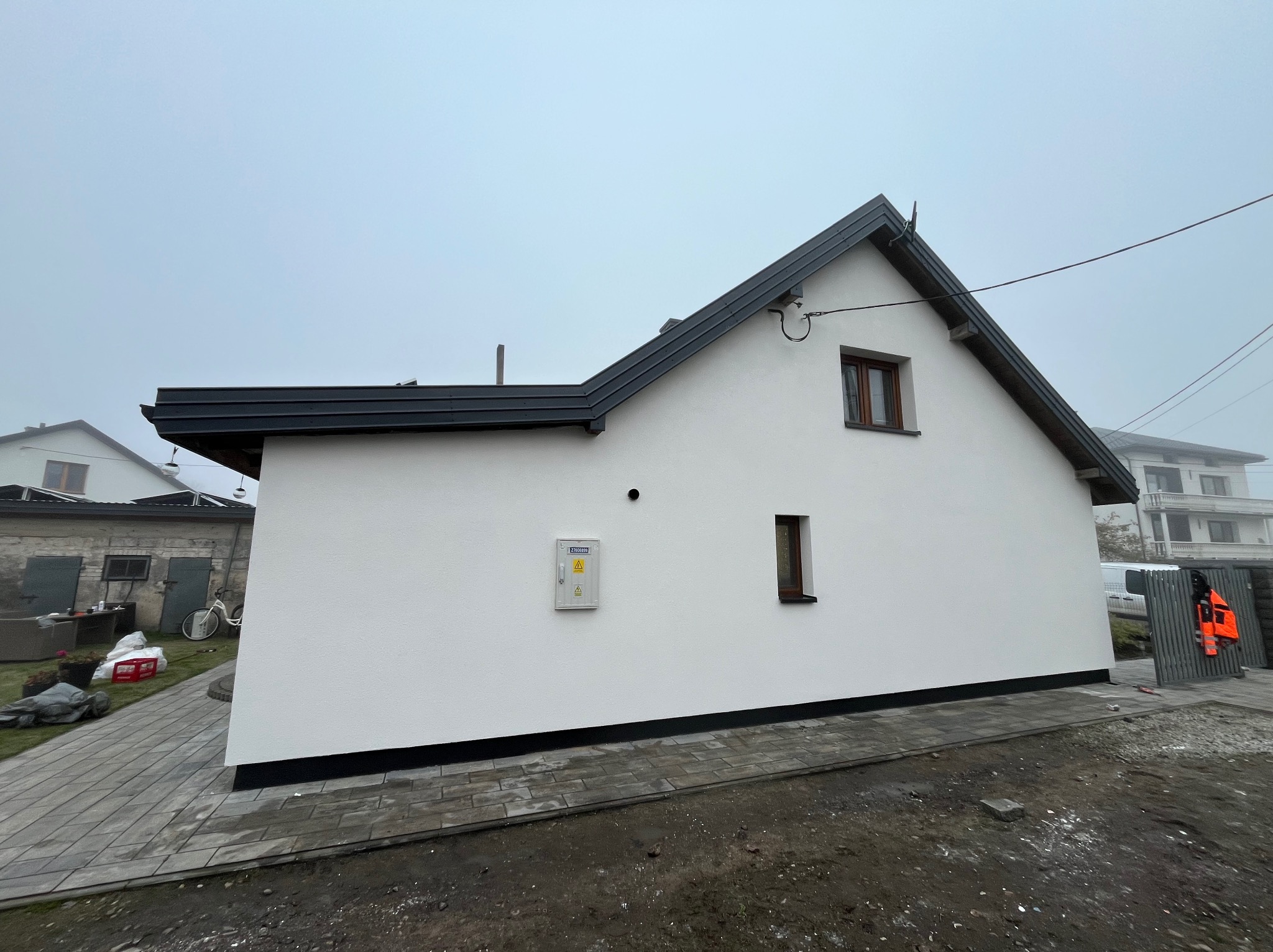 Otynkowany na biało budynek z ciemną dachówką i dwoma małymi oknami, widok z boku na tle pochmurnego nieba i sąsiednich budynków. Kostka brukowa wokół budynku.