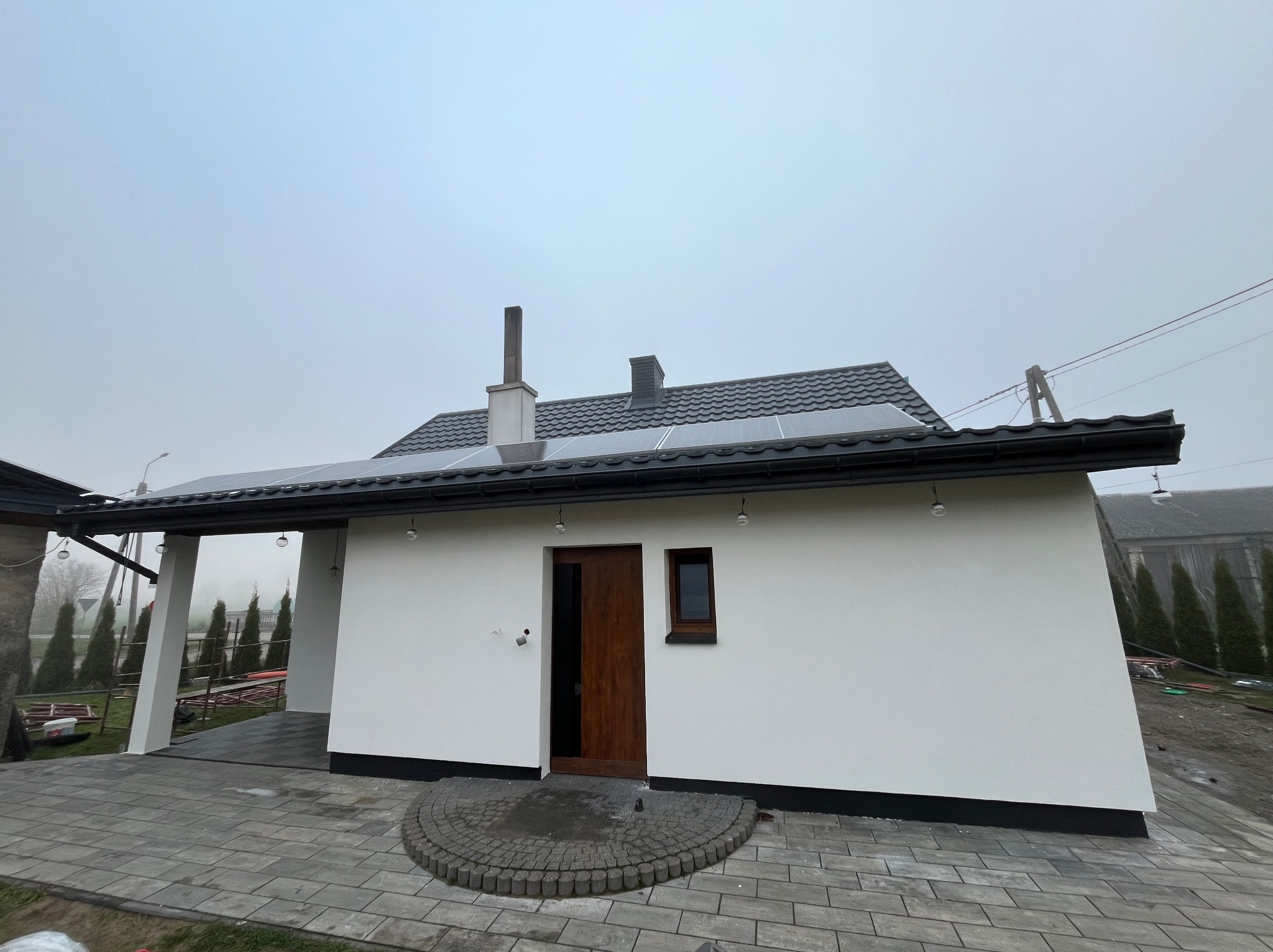 Biały dom z ciemną stolarką i panelami słonecznymi na dachu, widok z zewnątrz. Ułożona kostka brukowa przed wejściem. Mglista pogoda.