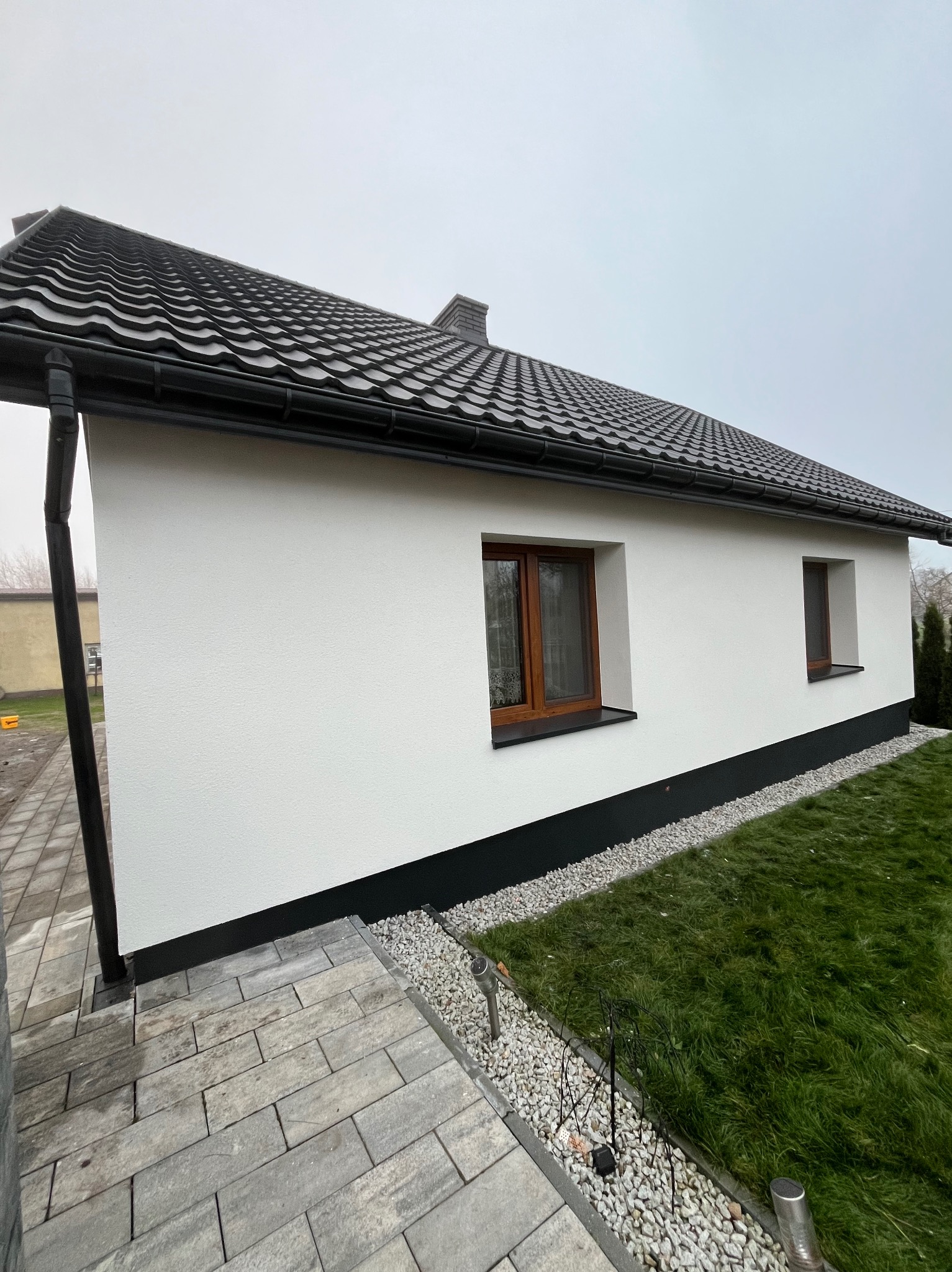 Biała elewacja domu jednorodzinnego z brązowymi oknami i ciemną podmurówką, widok z boku na tle szarej kostki brukowej i zielonego trawnika.