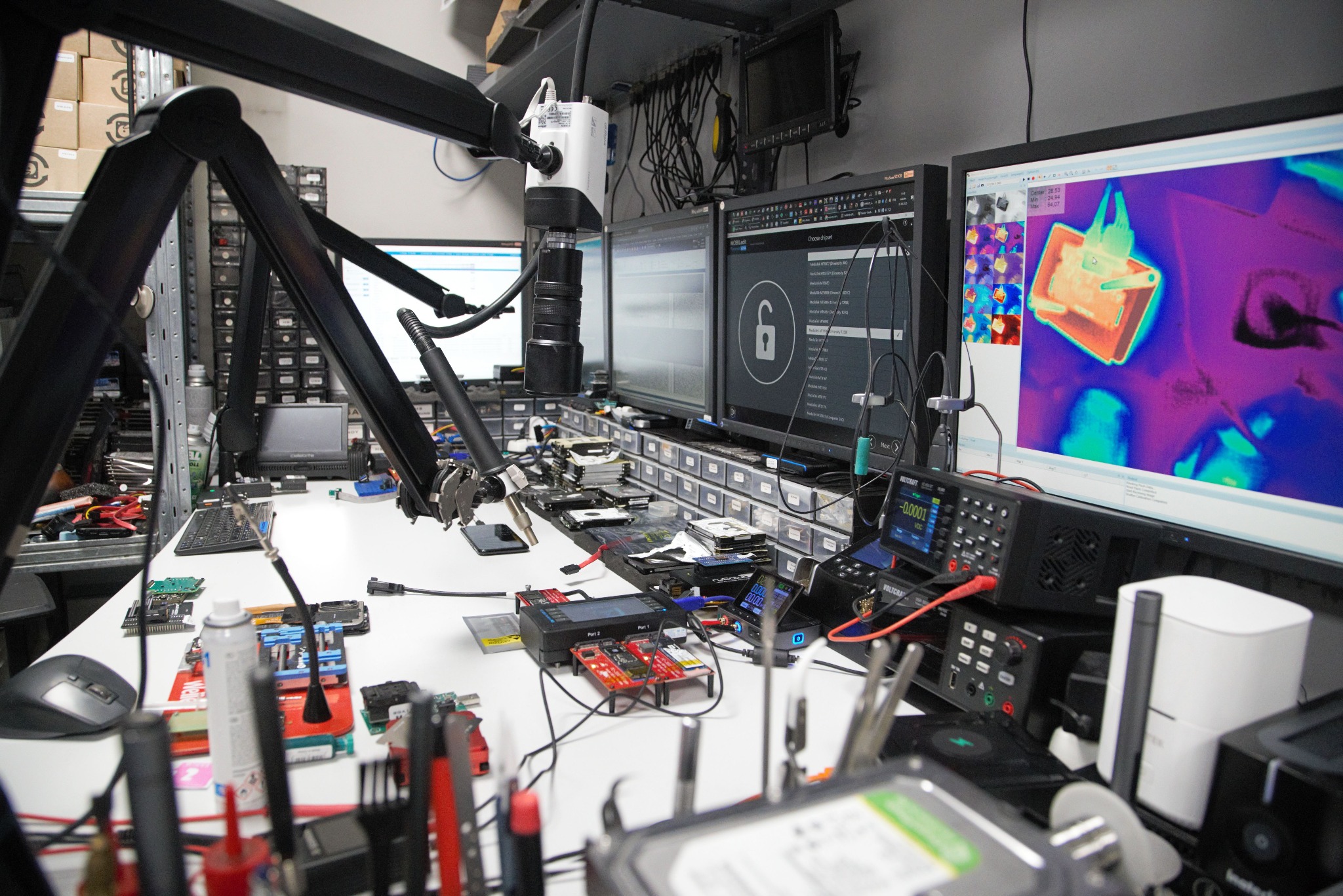Stanowisko pracy z elektroniką: monitory z programem do odzyskiwania danych, narzędzia, płyty główne, dyski twarde, mierniki i kamera inspekcyjna na ramieniu.