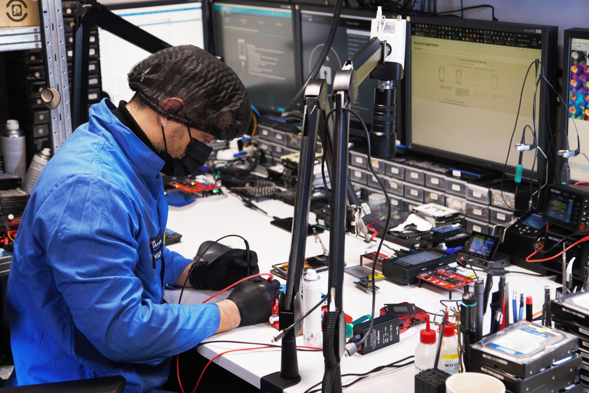 Technik w niebieskim kombinezonie i masce, z lupą, lutuje płytkę na stole pełnym sprzętu elektronicznego i dysków twardych w laboratorium IT.