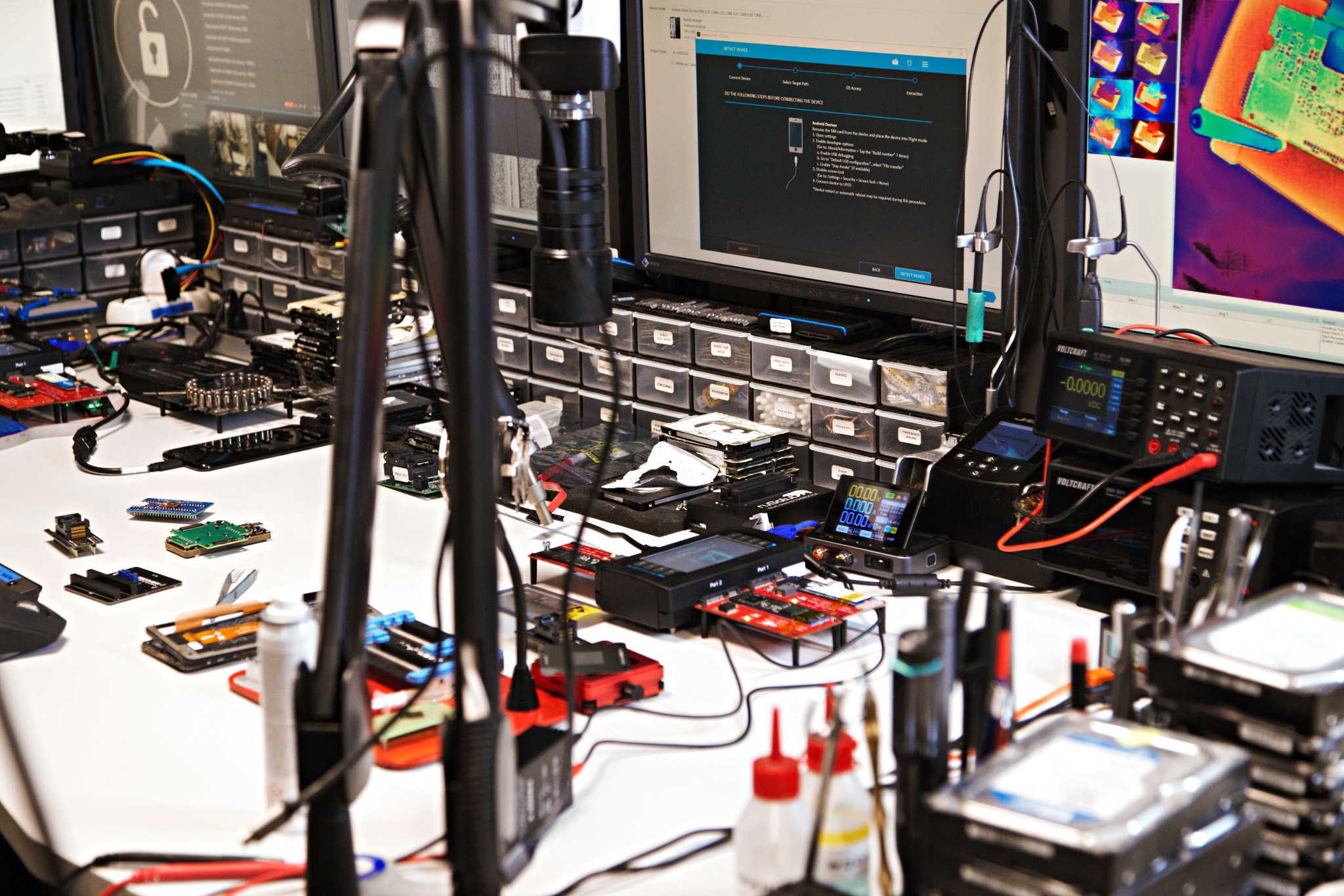 Stanowisko pracy z elektroniką, monitory i narzędzia do odzyskiwania danych. Widoczne dyski twarde, płyty główne i specjalistyczne urządzenia pomiarowe.