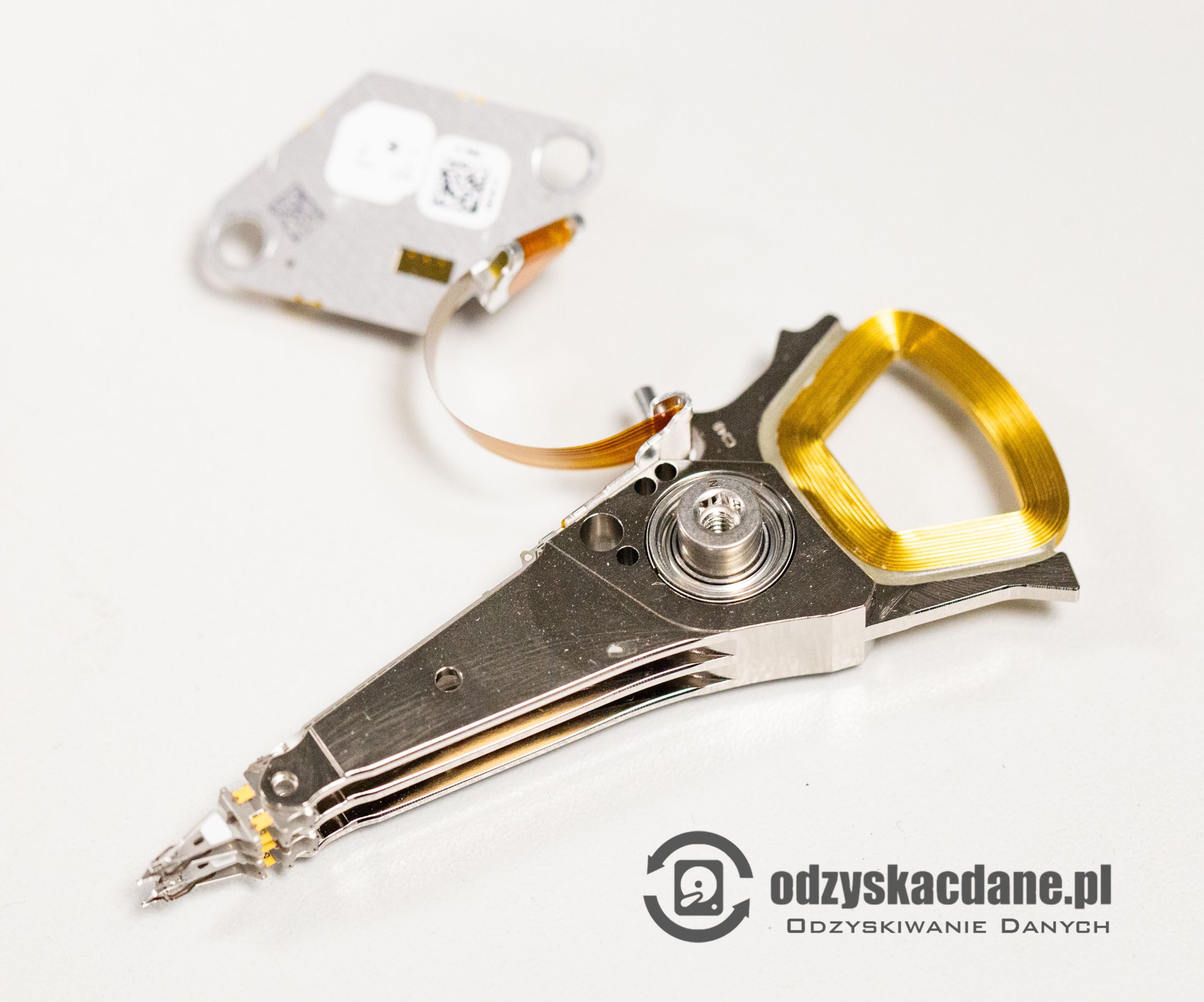 Głowica dysku twardego z elementami elektroniki, widoczne detale mechaniczne i złącza, na białym tle. Specjalistyczny sprzęt w procesie naprawy nośnika danych.