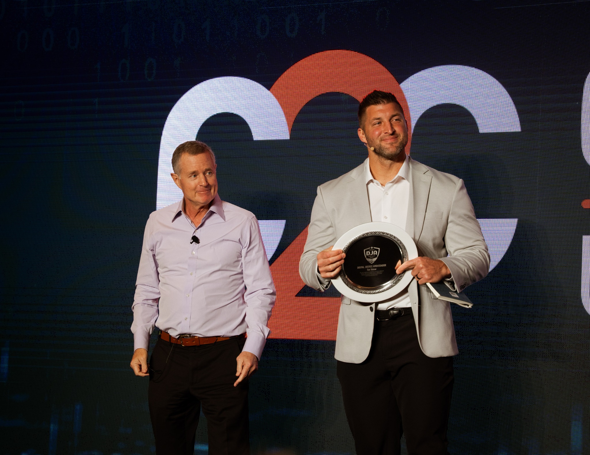 Dwóch mężczyzn na scenie: jeden trzyma nagrodę 'Digital Justice Ambassador', drugi z mikrofonem przypiętym do koszuli. Tło stanowi ekran z cyfrowym wzorem.