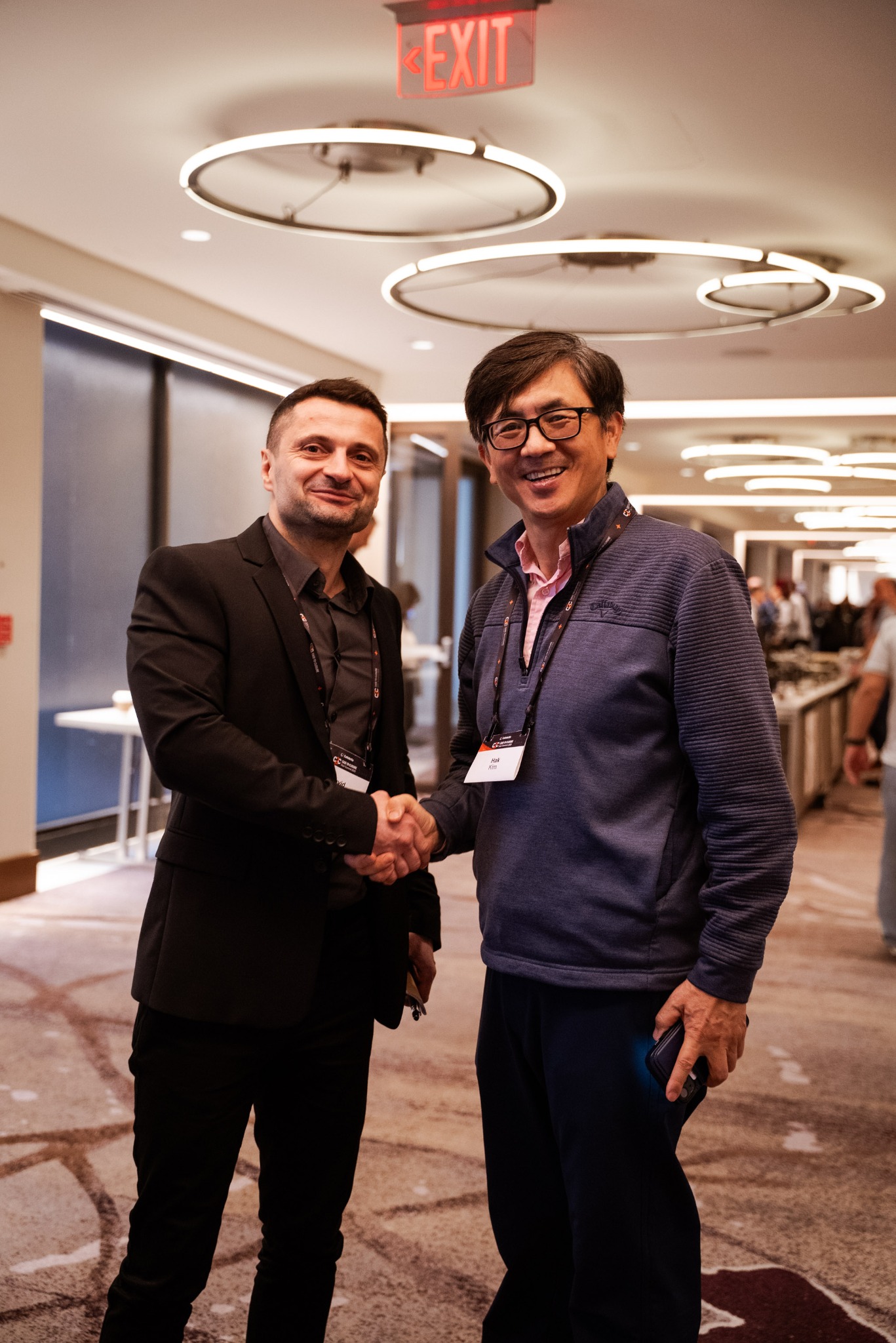 Dwóch mężczyzn w garniturach ściska dłonie na tle sali konferencyjnej z oświetleniem sufitowym i znakiem 'EXIT'. Widoczne identyfikatory konferencyjne.