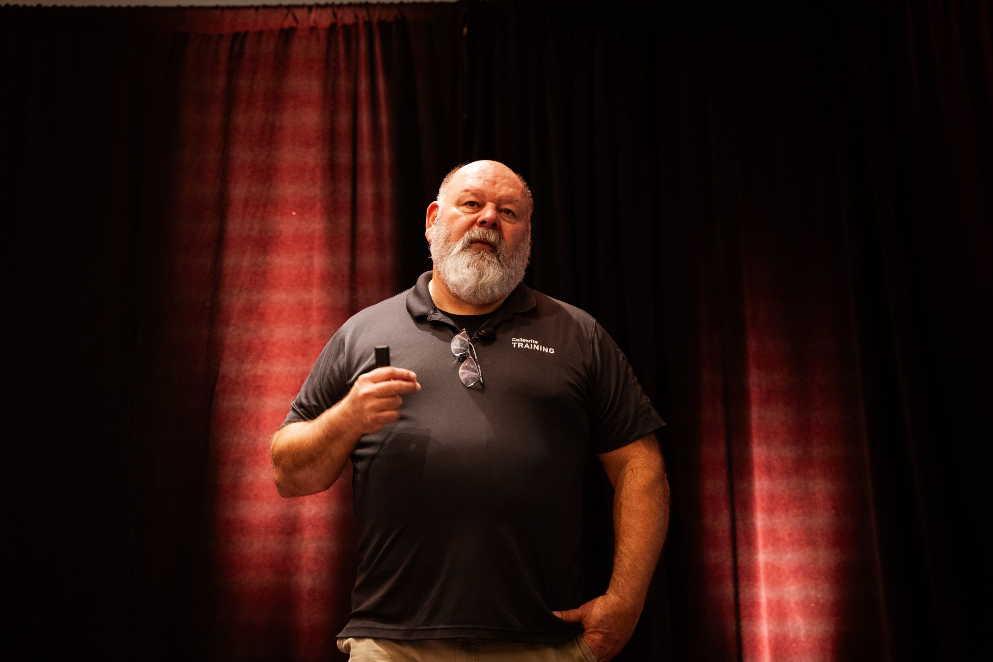 Mężczyzna z brodą w koszulce z napisem 'TRAINING' trzyma pendrive na tle czerwono-czarnych zasłon.